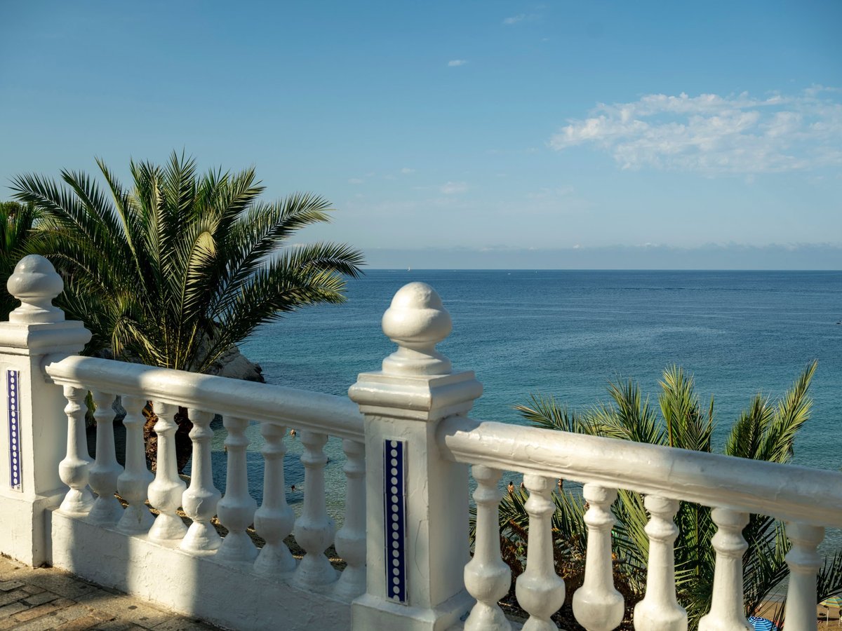 A classic white Mediterranean balustrade and palm fronds framing a vivid deep-blue Mediterranean sea with distant mountains on the horizon