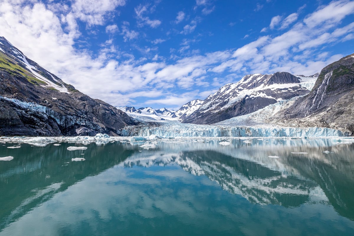 Massive tidewater glacier surrounded by snow-covered Alaskan peaks, reflected in the still turquoise water of a glacial fjord